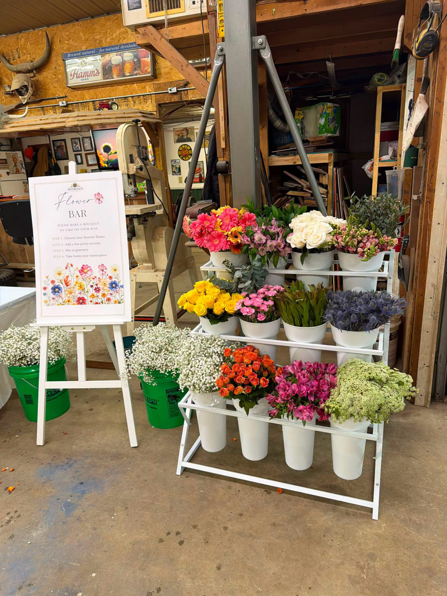 White tiered flower bar stand holding buckets of colorful blooms beside a make-a-bouquet sign